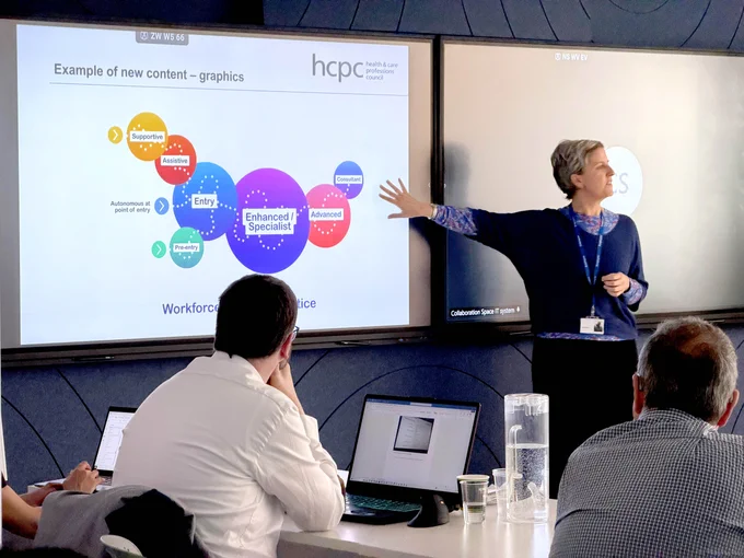 Presenter explaining a colorful graphic showing levels within the workforce on a large screen to seated attendees in a conference room.