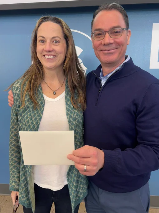 The Woman (Left): She has long, light brown hair and is smiling warmly at the camera. She wears a white t-shirt layered under a long, open-front green and grey patterned cardigan. She is also wearing a simple necklace with a dark pendant.

The Man (Right): He has short, dark hair and wears rectangular glasses. He is dressed in a navy blue quarter-zip sweater over a light-colored button-down shirt. He has his arm around the woman's shoulder and is holding a plain white envelope or card in front of them.