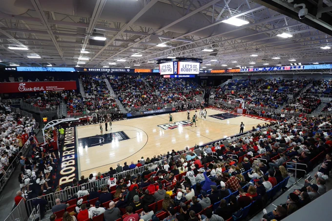 A basketball game is taking place in a large indoor stadium with a full audience. The court has "Robert Morris" written on it, and digital screens display messages related to the game. The venue is brightly lit, and the crowd is attentively watching the match.