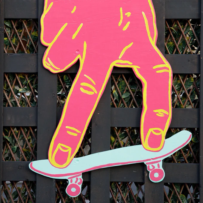 A colorful wooden cutout attached to a lattice wall depicts a hand performing a skateboarding trick on a miniature skateboard.
