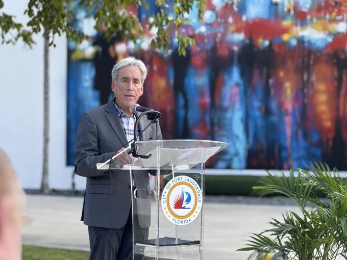 Man in a gray suit speaking at a clear podium with the City of Fort Lauderdale emblem outdoors near greenery.