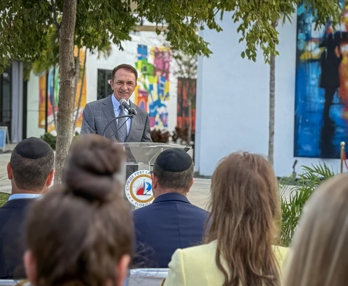 Speaker addressing an outdoor audience at a podium with a city emblem, surrounded by colorful murals and greenery.