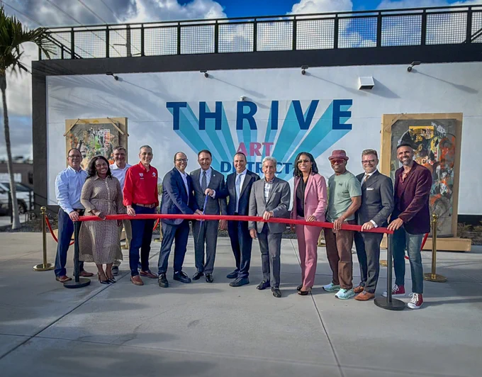 Group of diverse people in formal and casual attire holding a red ribbon for a grand opening event in front of a mural reading 'THRIVE ART DISTRICT'.