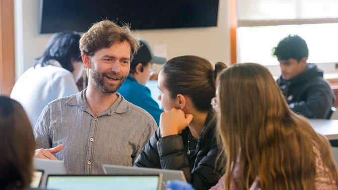 A male professor sits near students at a desk, their heads are turned to look at him as he attempts to explain a concept.