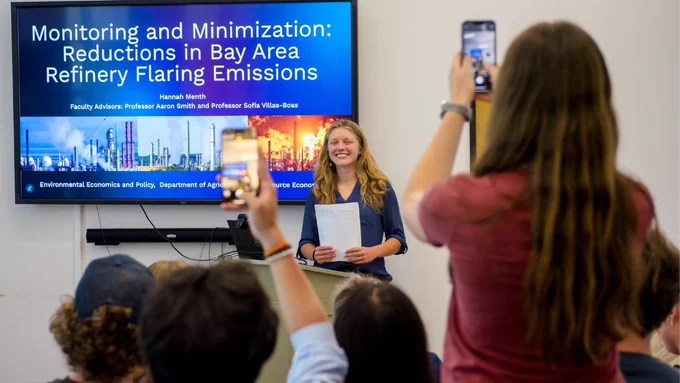 A photo of a young woman smiling as she stands at a podium. Several people stand in the foreground with their cell phones out and attempt to take photos.
