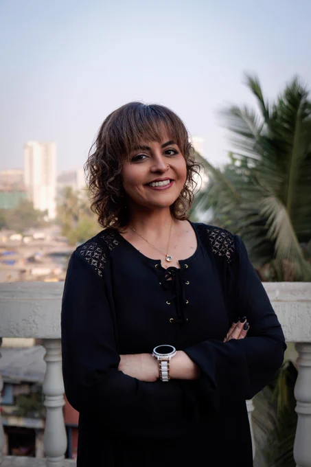 Ash is an Asian woman in her twenties with brunette shoulder-length hair in soft curls and a fringe. She stands on a rooftop overlooking a city also with palm trees.