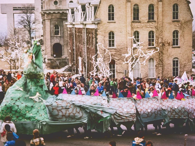 A large, colorful dragon float is paraded down a street by a group of people. The float is made of fabric and features scales and a long tail. In the background, there are historical buildings, on the Cornell University Arts Quad. Spectators are visible along the street.