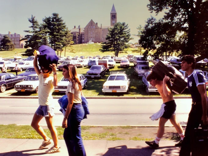 People carrying bags and belongings walk along a sidewalk on a sunny day. In the background, Cornell University's Uris Hall and its clock tower are visible, with a large expanse of parked cars and trees.