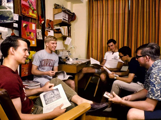 A group of people sit and engage in conversation inside a dorm room at Cornell University. The room is decorated with various posters and bookshelves filled with items. Some hold papers or books while others listen attentively. The setting suggests a casual and studious environment.