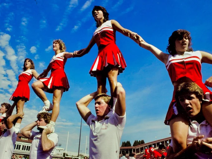Cheerleaders in red and white uniforms perform a formation outdoors at a Cornell University event. Bright blue sky with scattered clouds in the background.