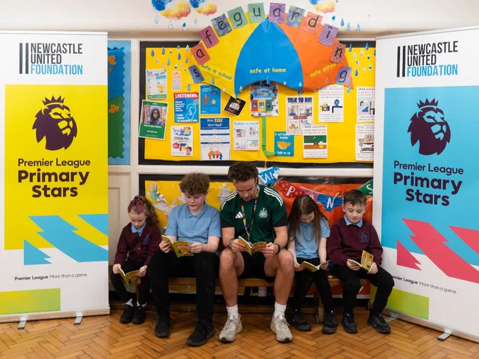 A group of children and an adult sitting and reading books together in a classroom. They are surrounded by colourful educational posters and banners for "Newcastle United Foundation" and "Premier League Primary Stars."