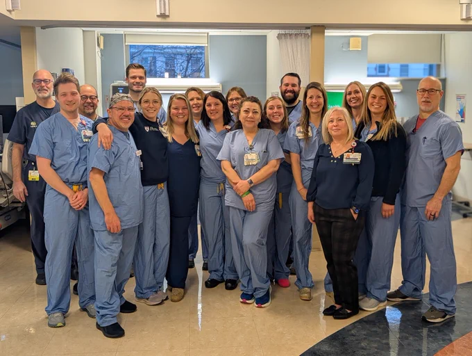 A group of about 18 healthcare workers stand in a hallway, posing for a photo. Most wear blue scrubs, with a few in dark tops. They are smiling, with a window and hallway visible in the background.