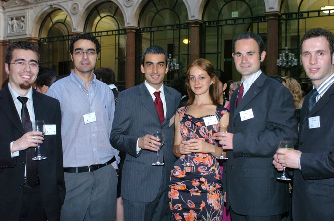 Group of six professionally dressed people holding drinks at an elegant indoor networking event with arched windows in the background.