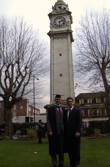 Two men in formal attire standing in front of a tall clock tower surrounded by leafless trees and buildings on a cloudy day.