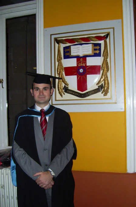 Graduate in cap and gown standing indoors beside a framed university crest on a yellow wall.