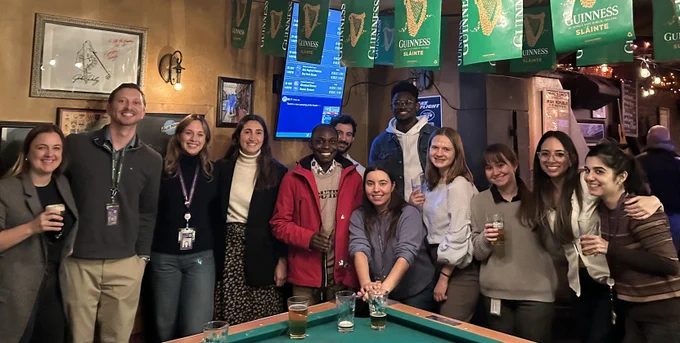 Group of friends posing with drinks around a pool table in a cozy pub decorated with Guinness banners.