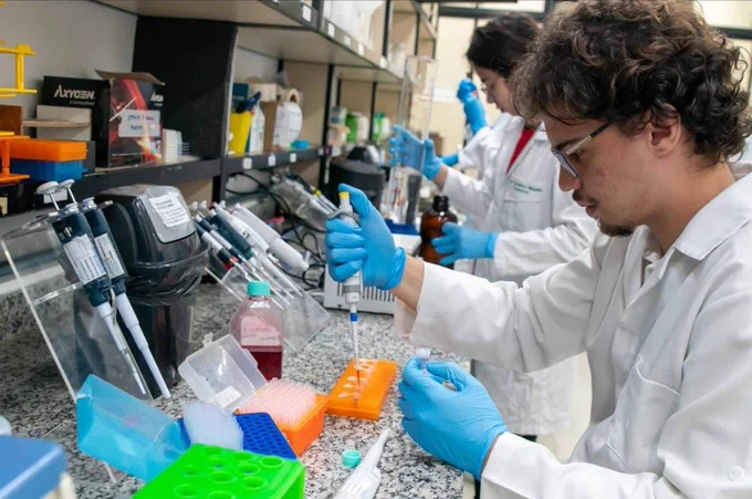 A imagem mostra um ambiente de laboratório. No primeiro plano, há um pesquisador usando jaleco branco, óculos de proteção e luvas azuis. Ele segura uma micropipeta e está transferindo líquido para pequenos tubos organizados em uma placa de microtubos laranja que está sobre uma bancada de granito. Nela, há diversos materiais típicos de laboratório. O foco dele está no procedimento que realiza.
