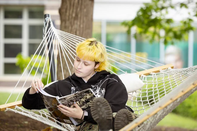 A person with bright yellow hair relaxes in a white rope hammock outdoors, leaning against a white pillow while reading a book or notebook. They wear a black hoodie with white graphics and camouflage pants. The hammock is tied between a tree and a metal pole, surrounded by green grass and trees, with a building featuring large windows in the background. The scene feels calm and studious, set in a quiet outdoor campus space.