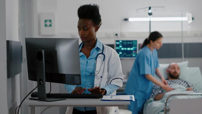 A healthcare professional in a white coat and stethoscope stands at a computer station, typing. In the background, a nurse in scrubs tends to a patient lying on a hospital bed, with medical equipment visible.