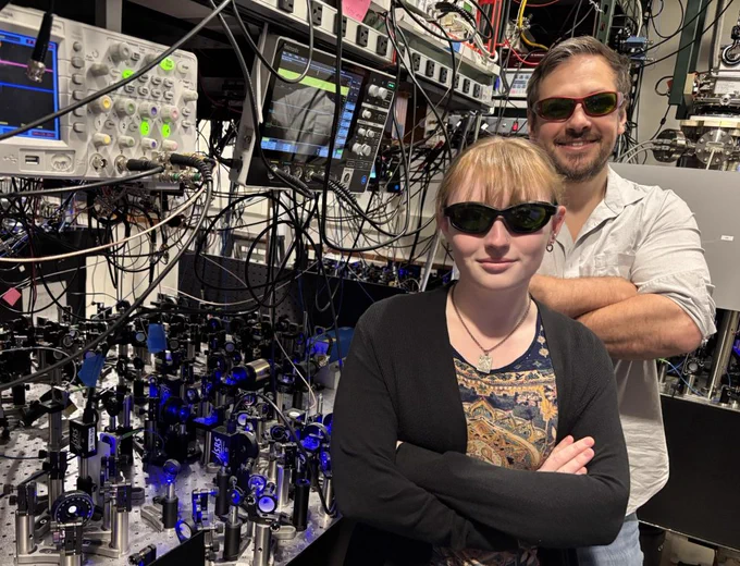 Two researchers stand in a lab filled with complex optical equipment and numerous cables connected to oscilloscopes and monitors.