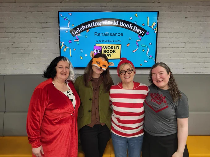 A group of four people standing and smiling in front of a screen displaying "Celebrating World Book Day" with colorful confetti graphics. They are wearing casual and festive attire, with one person wearing a fox mask. The screen mentions "Renaissance" and features the "World Book Day" logo.