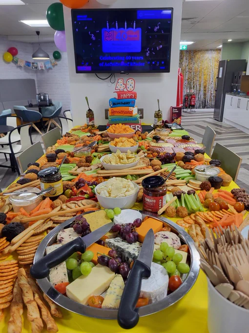 A long table is filled with an assortment of snacks, including cheese, grapes, crackers, vegetables, and dips. Knives are placed on cheese platters. A digital screen in the background celebrates "40 years of Andrew's" with festive balloons nearby. The setting appears to be in a decorated dining area.