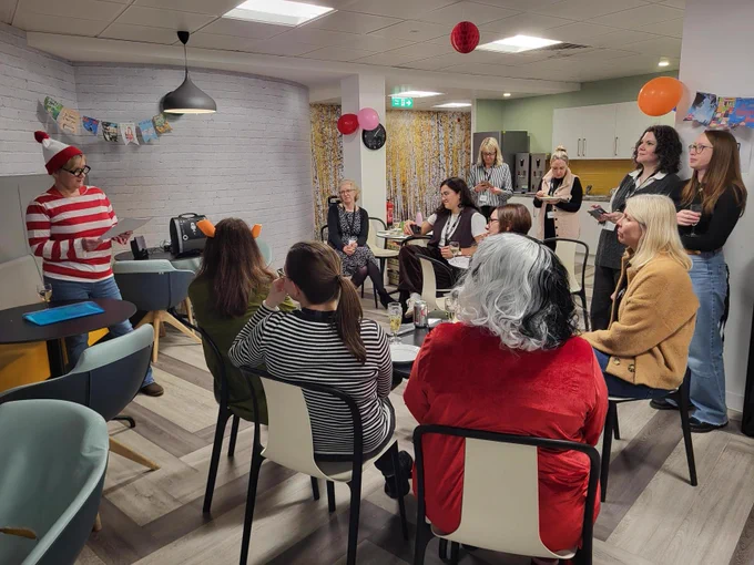 A group of people sitting and standing in a decorated room, engaging in conversation. Some are in costume, including a person dressed as Waldo from "Where's Waldo?". The room has festive decorations and a dining area setup.