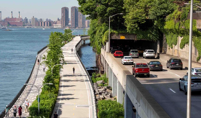 A riverside walkway lined with trees and lampposts runs parallel to a busy road entering a tunnel beneath a green hillside.