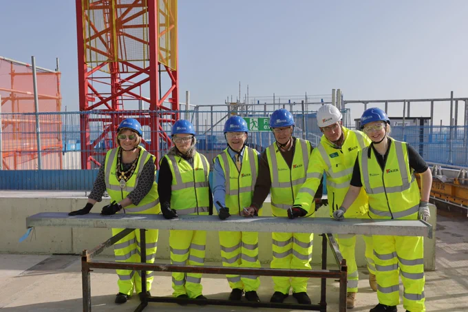 A group of six people wearing high-visibility vests, hard hats, and gloves are standing together at a construction site, smiling for the camera. They are gathered around a metal beam, with a tall red and yellow crane structure visible in the background. Blue fencing surrounds the site under a clear sky.