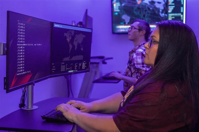 Two individuals are working at computer stations in a dimly lit room with purple lighting. The first person is focused on a large monitor displaying code and a digital map. In the background, another person is engaging with multiple screens, one of which shows detailed data and maps. The setting appears to be a tech environment, possibly related to cybersecurity.