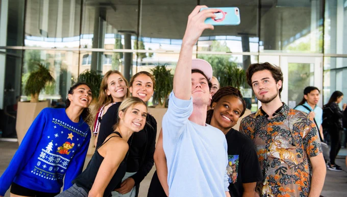 Group of young adults posing for a selfie outside a modern building with large glass windows.