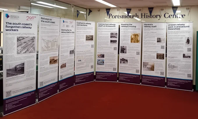 Eight pull-up banners, from the 'South coast's forgotten railway workers' exhibition, stood on a red carpet, around the edges of a room. The banners introduce railway work before 1939 in the Portsmouth area, and some of the employees' life stories. Above the banners and behind, a sign says 'Portsmouth History Centre', with 'Portsmouth 100' bunting on the ceiling.