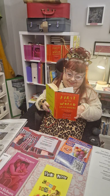 Penny sits in front of her Living Archive and books for sale, which are on white shelving behind her to the left. The books we can see are Desires Reborn - a pink paperback with white lettering and a faint image of a disabled couple embracing, next to this is the hardback memoir First In The World Somewhere, it is a red cover with bright yellow lettering and white text. In the lower third there is a graphic of lips outlined in black. 
Below this shelf are many files and other books featuring Penny’s work. On top of the shelving there are two vintage suitcases, to the right of the image there’s a framed picture of Penny’s 1989 seven inch single, a black and white chalk effect image of a seated girl. Below this is another framed picture announcing Penny’s Living Archive. In the centre, Penny sits in her wheelchair. She wears glasses,  has red hair, up in a bun and is wearing a headband made of twigs and crystals into the shape of antlers. Penny is wearing a leopard print dress and a crop