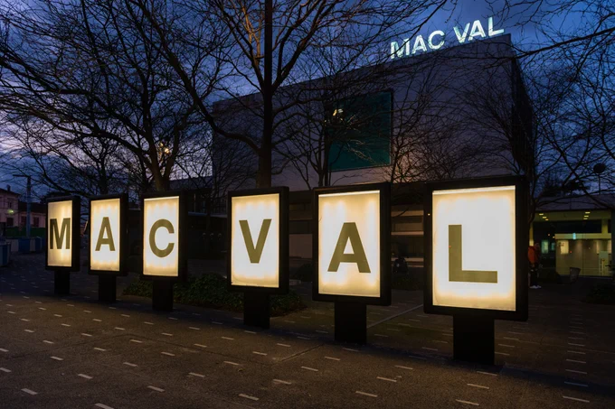 Vue nocturne du musée MAC VAL à Vitry-sur-Seine, avec de grands panneaux lumineux affichant chaque lettre du nom « MAC VAL » devant le bâtiment moderne, des arbres sans feuilles se découpant sur le ciel bleu sombre en arrière-plan.