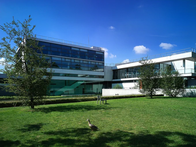Bâtiment moderne en verre et béton situé derrière une pelouse verte bien entretenue, avec quelques arbres et un banc. Un canard marche sur l'herbe au premier plan, sous un ciel bleu parsemé de nuages blancs.