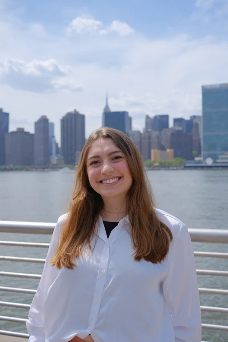 Melanie, a white woman with long brown hair, stands in front of the river in New York City, smiling. 