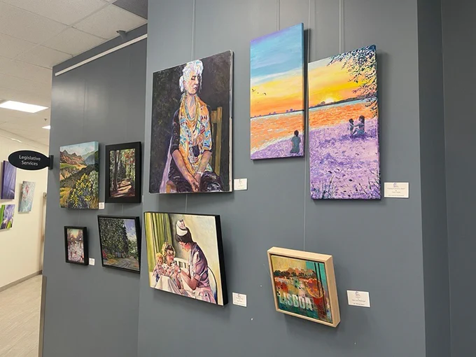 This image shows a wall with various colorful paintings on display in an exhibition space. Notable works include landscapes, a portrait of a seated person, and a painting of people by a sunset-lit ocean. The wall is labeled "Legislative Services.