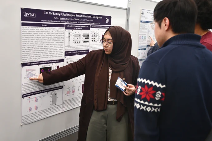 A student presents a poster titled “The Cbl Family Ubiquitin Ligases Regulate Directional T Cell Migration” while pointing to a diagram and explaining the research to another attendee.