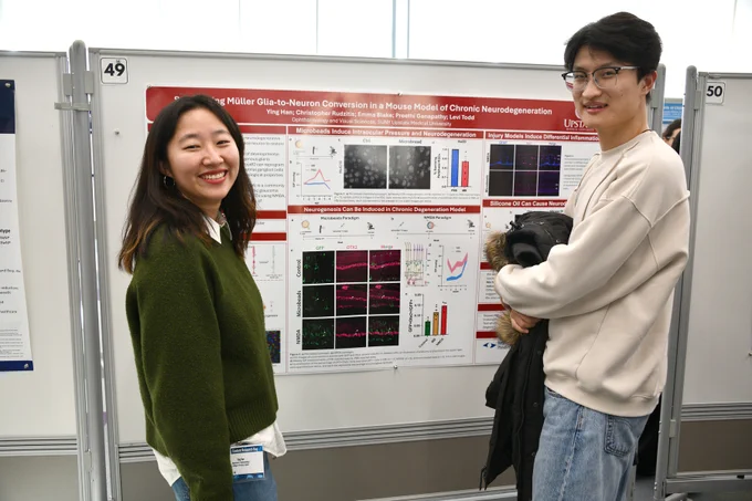 Two student researchers smile while standing beside their poster titled “Stimulating Müller Glia-to-Neuron Conversion in a Mouse Model of Chronic Neurodegeneration” at Upstate’s Student Research Day.