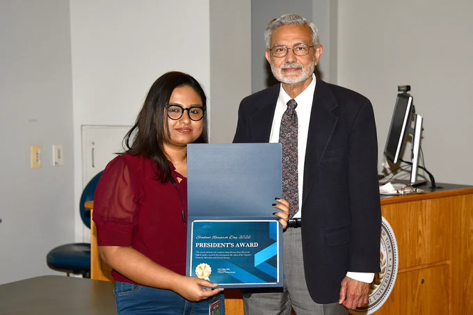 A student holds a plaque reading “Student Research Day 2026 – President’s Award” while standing beside Upstate President Dr. Dewan at a podium inside a lecture hall at SUNY Upstate Medical University.