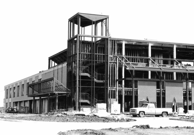 A black and white image of a partially constructed building with steel framework visible. The structure is surrounded by dirt grounds and construction materials. A pickup truck is parked nearby.