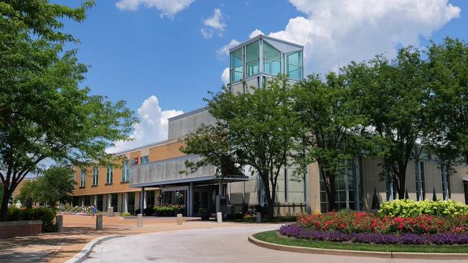 A large modern building with a glass tower section is surrounded by lush green trees and colorful flower beds. The sky is bright blue with scattered clouds, creating a serene atmosphere.