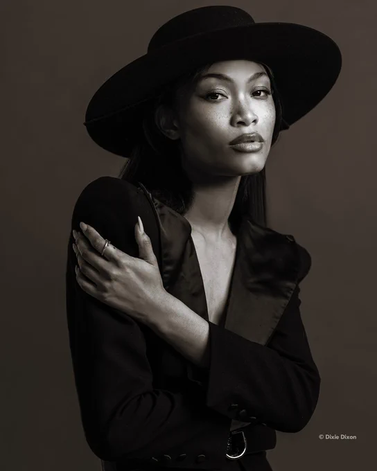 A photo of a woman with a calm, direct gaze wearing a wide-brim hat and dark clothing, posed against a plain background, captured in black and white. Text in the lower right-hand corner reads:\u201C\u00A9 Dixie Dixon.\u201D