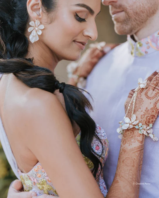 A photo of a woman in close embrace with a man, highlighting detailed henna designs on her arm, floral jewelry, and soft natural light during an intimate moment. Her dark hair rest on her left shoulder over a pastel-colored beaded dress. Text in the lower right-hand corner reads: \u201C\u00A9 Charmi Pe\u00F1a.\u201D
