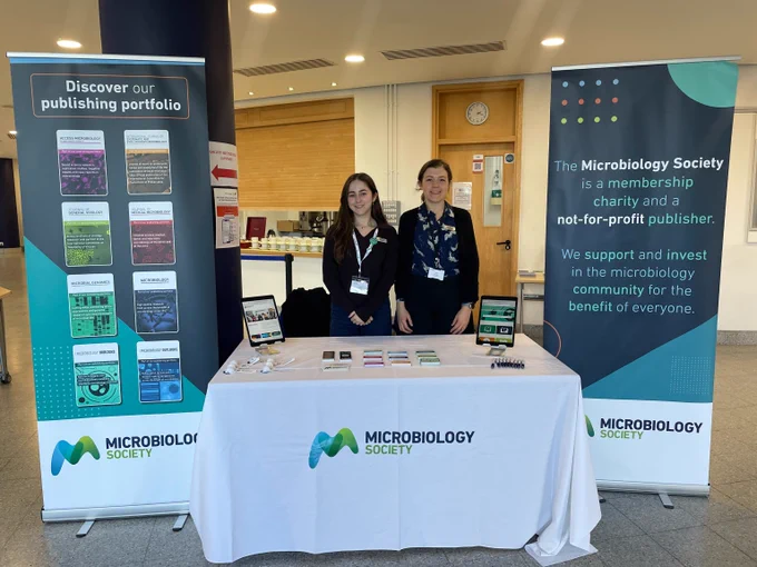 Two representatives stand behind a Microbiology Society booth with informational banners and promotional materials on display.