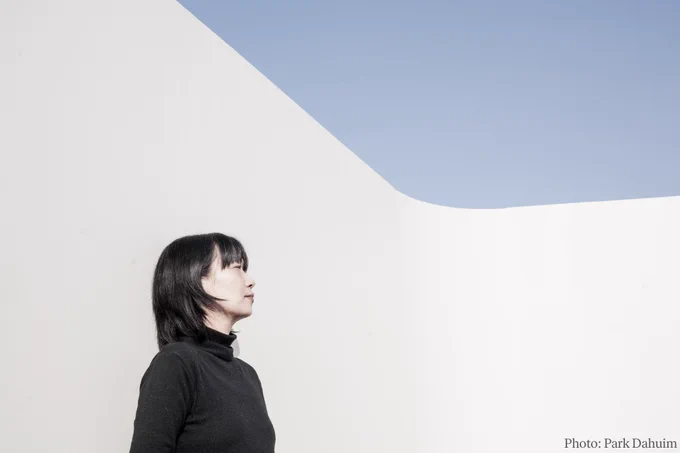 Han Kang stands against a minimalistic, abstract background of sharp geometric shapes, under a clear sky, peacefully looking to the side.