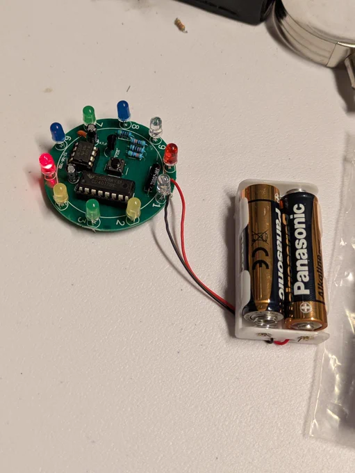 Close-up of a small round circuit board with multicolour LEDs lit up, connected by wires to a two-AA battery holder.