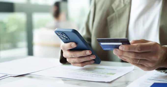 Close-up of a person holding a smartphone in one hand and a bank card in the other while looking at paperwork on a table, suggesting an online payment or banking task.