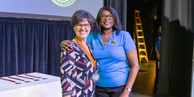 Two individuals stand together on a stage, smiling. One is wearing a patterned outfit with a medal; the other is in a blue shirt.
