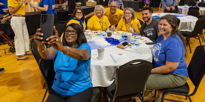 A group of faculty and staff are gathered around a table at an event, smiling and posing for a selfie with Dr. Williams. The setting appears festive, with lighted decorations and other attendees in the background.